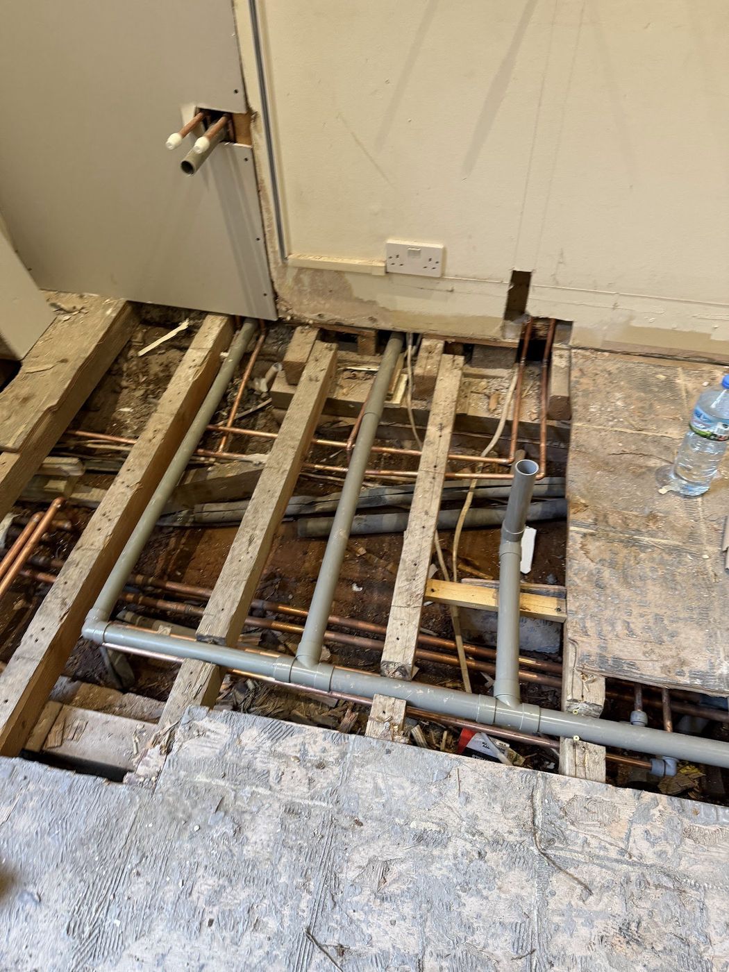 Floorboards lifted to expose copper and waste pipe runs between joists
